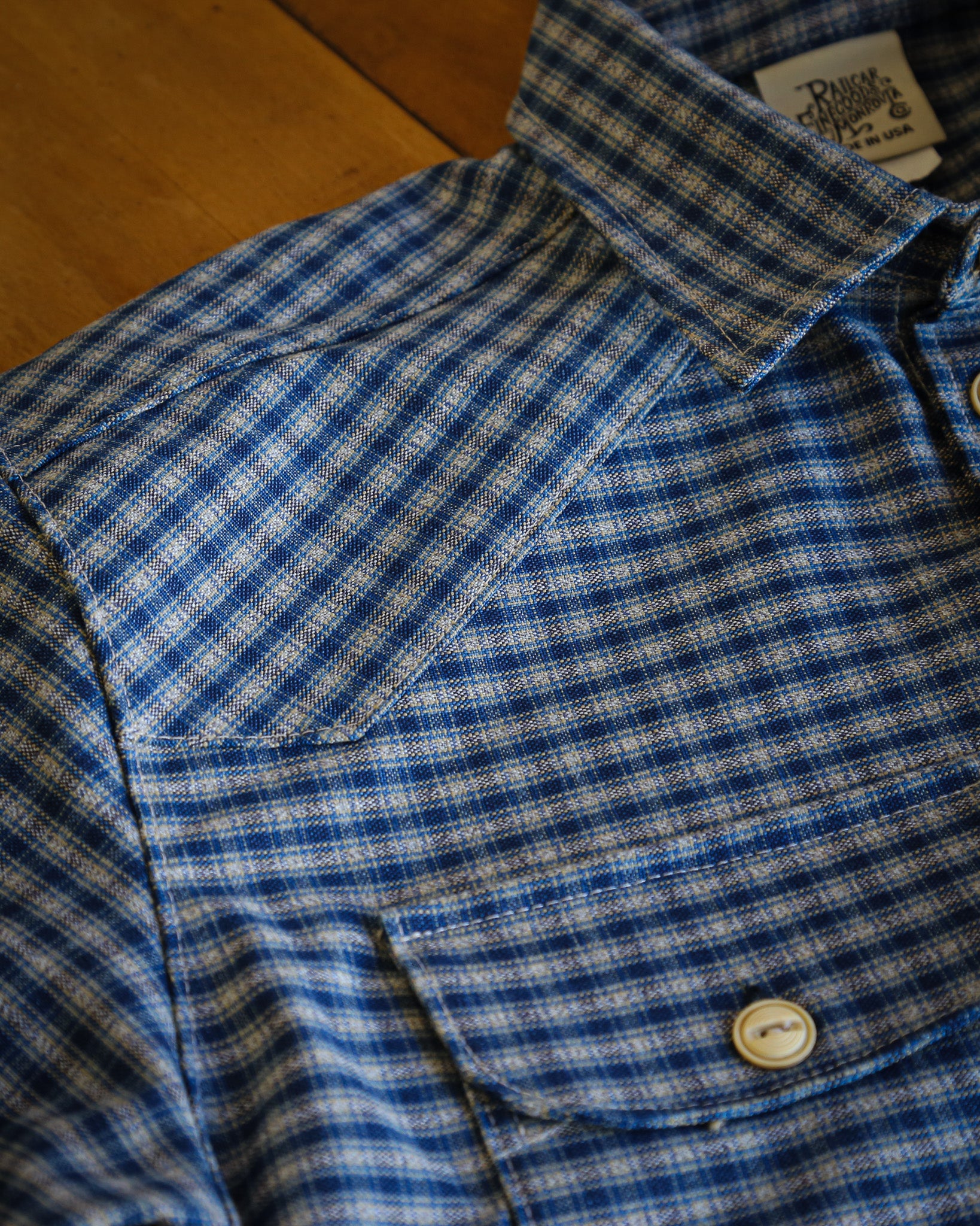 Close-up of a blue and white checkered shirt with a button and tag on a wooden surface.