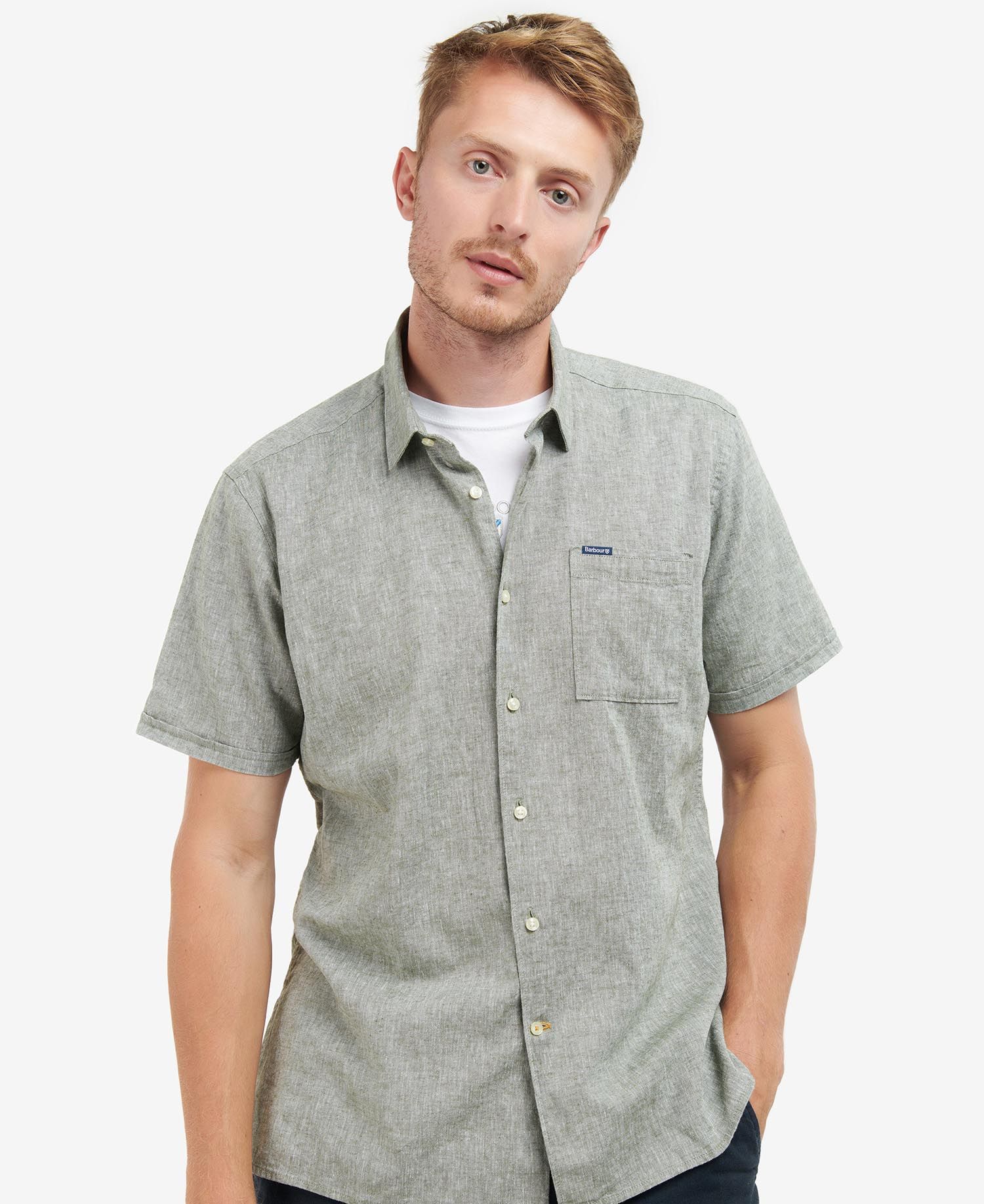 A male model wearing a short-sleeved, button-up Barbour Nelson summer shirt in a bleached olive color with a left chest pocket and visible Barbour and Shield label.